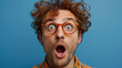 Surprised young man with curly hair and round glasses, mouth open in shock against blue background.