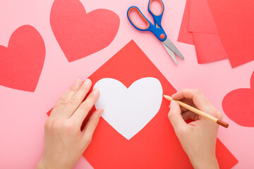 Wall Mural - Young adult woman hand outlining contour with pencil for cutting red heart shape from application paper on pastel pink table background. Making decoration. Closeup. Point of view shot. Top down view.