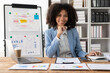 © ArLawKa - Cheerful businesswoman sitting at desk and looking at camera, African American executive smiling using laptop while working from office with financial graph document for financial business analysis.