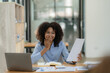 © ArLawKa - Cheerful businesswoman sitting at desk and looking at camera, African American executive smiling using laptop while working from office with financial graph document for financial business analysis.