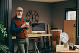 Fototapeta  - Portrait d'un homme debout souriant quinquagénaire senior hipster élégant et stylé qui fait une pause et qui boit un grand café dans un atelier créatif vintage