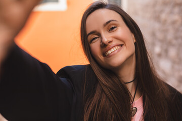 Sticker - Close up portrait of young widely smiling woman tourist posing, walking in old town. Happy young model makes photo holding camera in hand close-up. Outdoor selfie photo in fashion outfit while walking