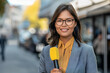 A professional asian journalist wearing glasses holds a yellow microphone in front of her and reports the news