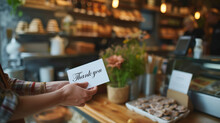 A Woman Is Holding A Thank You Card In Front Of A Bakery. The Card Is White And Has A Black Message That Reads "Thank You." The Woman Is Standing In Front Of A Counter With Various Baked Goods