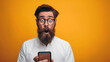 Photo of a 23 year old man with beard in a white shirt wearing glasses with a shocked expression while looking at a cellphone screen, studio photo