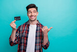 Fototapeta Panele - Photo of cheerful dreamy man dressed plaid shirt rising bank card pointing thumb empty space isolated turquoise blue color background