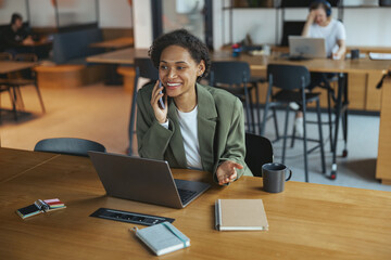 Wall Mural - A woman on a chair at a wooden desk, using a laptop, talking on the phone in nice modern coworking office