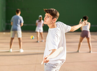 Wall Mural - Portrait of sporty young guy playing pelota on open fronton court on summer day, ready to hit rubber ball with traditional wooden racket. Sport and active lifestyle concept