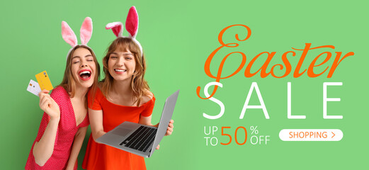 Poster - Happy young women in bunny ears headbands holding laptop and credit cards on green background. Online shopping for Easter