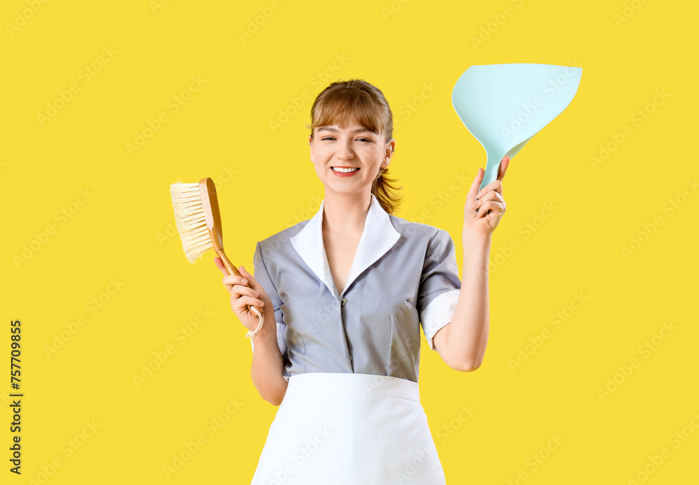 Young chambermaid with dustpan and brush on yellow background