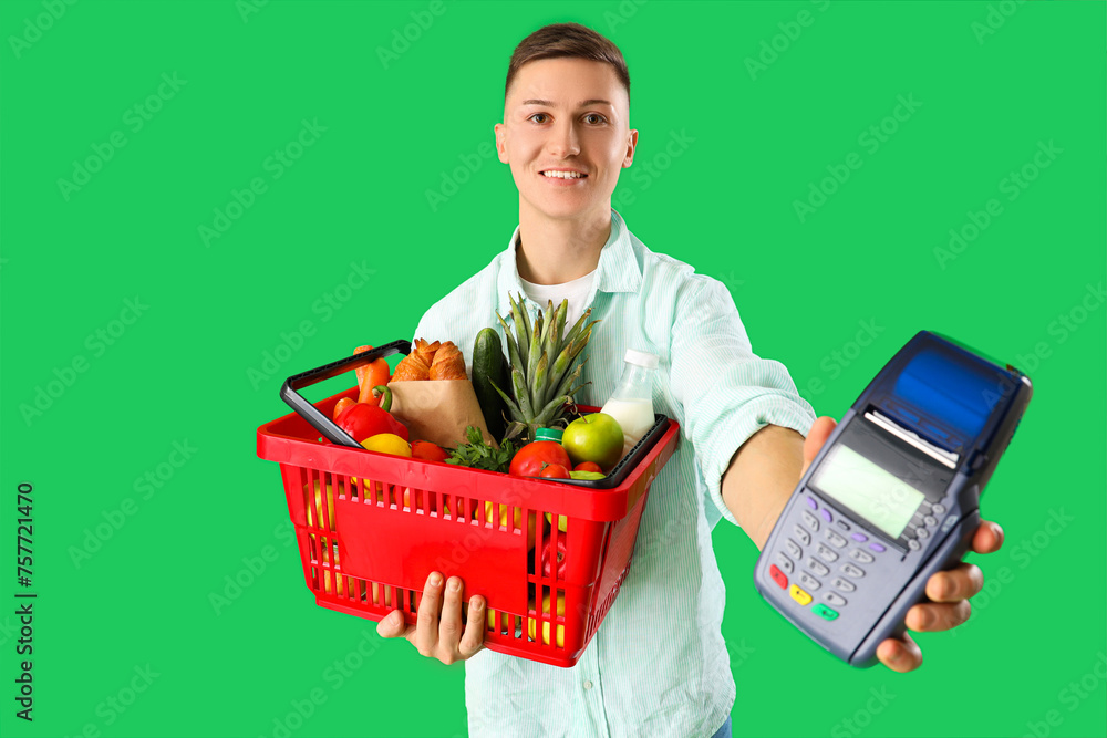 Young man with full shopping basket and payment terminal on green background