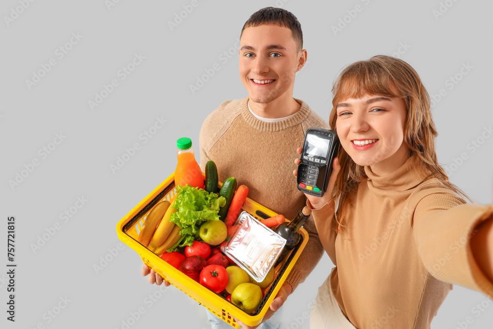 Young couple with full shopping basket and payment terminal taking selfie on light background