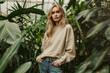 Portrait of an attractive blonde hair female model wearing beige color oversized crewneck blank mockup sweatshirt posing in a greenhouse with hand in pocket