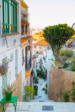 Fototapeta Miasto - Narrow street with steps, white houses and blue potted plants in ancient neighborhood Santa Cruz in Alicante old town on hillside. Costa Blanca on Mediterranean sea coast, Spain