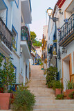 Fototapeta Miasto - Narrow street with steps, white houses and blue potted plants in ancient neighborhood Santa Cruz in Alicante old town on hillside. Costa Blanca on Mediterranean sea coast, Spain