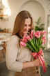 Beautiful woman is smelling a bunch of spring flowers and standing in living room at home. Female. International Women's Day. Portrait young girl in dress holding in hands a big bouquet of pink tulips