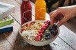 Female hands pick up a blueberry  from a Smoothie açai bowl with banana, strawberry, coconut flakes, chia, nuts, beautifully decorated. Served with bottle of juice on a wooden table. 