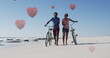 Image of hearts over happy african american couple walking with bikes on beach
