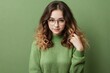 Young beautiful introverted woman with curly long hair, wearing a green sweater and glasses posing on a natural green background with copy space.