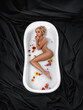 Beautiful woman with perfect legs and hips posing in the white bath with milk and flowers on a black fabric background. Rejuvenation skin care therapy.