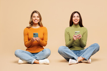 Wall Mural - Full body young friends two women they wear orange green shirt casual clothes together sit hold in hand use mobile cell phone isolated on plain pastel light beige background studio. Lifestyle concept.