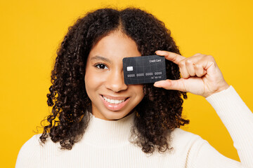 Little kid teen girl of African American ethnicity wears white casual clothes hold cover eye with mock up of credit bank card isolated on plain yellow background studio. Childhood lifestyle concept.