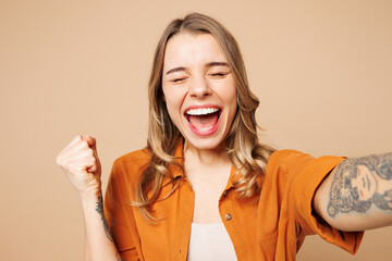 Wall Mural - Close up young woman wear orange shirt casual clothes doing selfie shot pov on mobile cell phone do winner gesture close eyes isolated on plain pastel light beige background studio. Lifestyle concept.