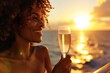 A close-up of a smiling black woman, holding a champagne glass and enjoying a sunset on the deck of a luxury cruise ship