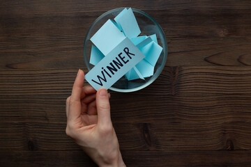 Female hand holding lottery ticket with winner word near glass bowl full of paper sheets. Winner concept