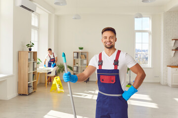 Wall Mural - Portrait of happy smiling man janitor from professional cleaning service in uniform looking cheerful at camera holding mop in hand mopping house or office. Housework and housekeeping concept.