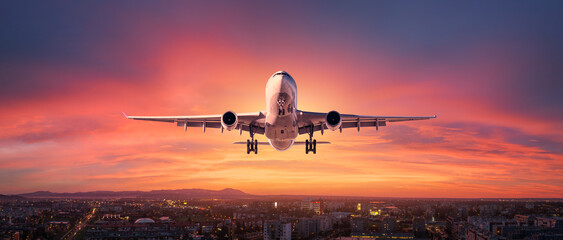 Airplane is flying in colorful sky over city at sunset. Landscape with passenger airplane, skyline, purple sky with red and pink clouds at dusk. Aircraft is landing at twilight. Aerial view of plane