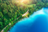 Fototapeta Kosmos - Aerial view of road near blue lake, green forest at sunrise in summer. Bled lake, Slovenia. Travel. Top view of beautiful road, trees in spring. Landscape with highway and sea bay. Road trip. Nature