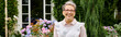 mature cheerful woman with glasses posing next to flowers and smiling happily at camera, banner