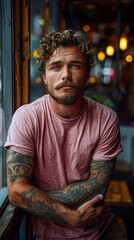 Wall Mural - Man with curly hair and tattoos is wearing a pink shirt, looking contemplatively at the camera