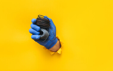 Wall Mural - A left man's hand in a blue knitted glove holds a two coil of steel wire. Torn hole in yellow paper. The concept of a worker, a labor migrant, a master of his craft. Copy space.