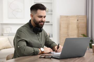 Poster - E-learning. Young man taking notes during online lesson at wooden table indoors