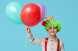 Funny little boy in clown wig and hat holding balloons on blue background. April Fools Day celebration
