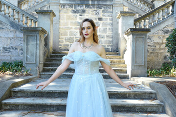 Wall Mural - Close up  portrait of beautiful female model wearing blue fantasy ballgown, like a fairytale elf princess.  Elegant pose, gestural hands reaching out, on the terrace of a romantic castle staircase 