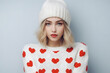 A young blonde woman wearing a white beanie and a sweater with red hearts gazes at the camera with a neutral expression