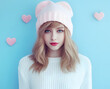 A young woman with a pink beanie hat and blue eyes on a blue background with pink heart shapes