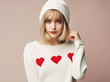 A stylish young blonde woman wearing a white beanie and a white sweater adorned with red hearts poses with a hand to her chin