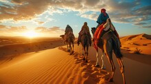 A caravan of camels with riders trek across rolling desert dunes under a vibrant sunset sky.