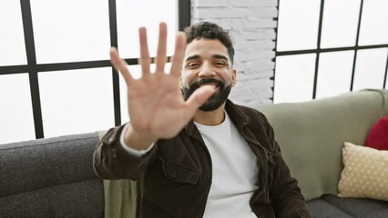 Wall Mural - Cheerful young man playfully pointing up with five fingers, showing off his confident smile at home