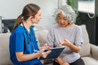 © NanSan - Caucasian teenager female doctor consults with and Asian elderly patient on medical history, discussing headaches, chest pain, coughing, sneezing, stomach pain, and neck pain while seated on a sofa.