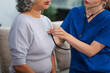© NanSan - Caucasian female doctor uses stethoscope to check heart rate on elderly Asian patient on sofa.