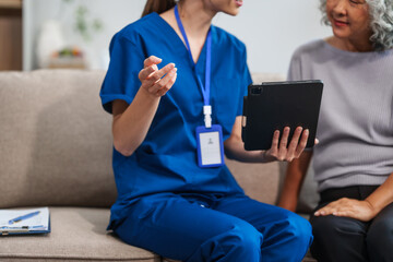 Caucasian female doctor conducts a check-up using a tablet device for an elderly Asian patient seated together on the sofa.