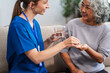 © NanSan - Caucasian female doctor provides a glass of water and medication pills to an elderly Asian patient while they are seated together on the sofa.
