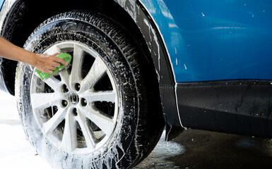 Wall Mural - A person is washing a car tire with a green brush. Tire is covered in water and soap. Blue car wash with white soap foam. Auto care service. Car cleaning service concept. Vehicle cleaning service.