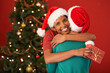 © M S/peopleimages.com - Women, hug and Christmas with surprise, gift box and package on red studio background. Face, people or friends with Xmas present or parcel with embrace or excited with celebration or bonding together