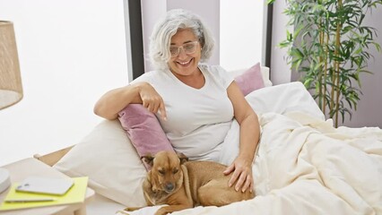 Wall Mural - Cheerful middle age woman with grey hair, sitting in her bedroom on the bed with her beloved dog, both wearing pyjamas. she's confidently pointing, showing something with a happy, beaming smile.